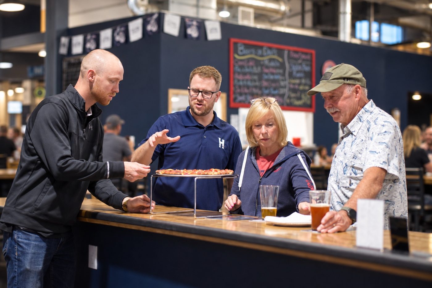 An owner serving a fresh pizza to an older couple and a guest standing at the bar
