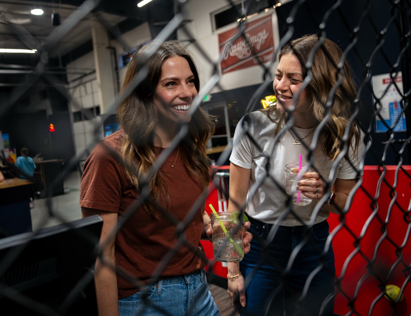 Two friends laughing together by the batting cages, drinks in hand